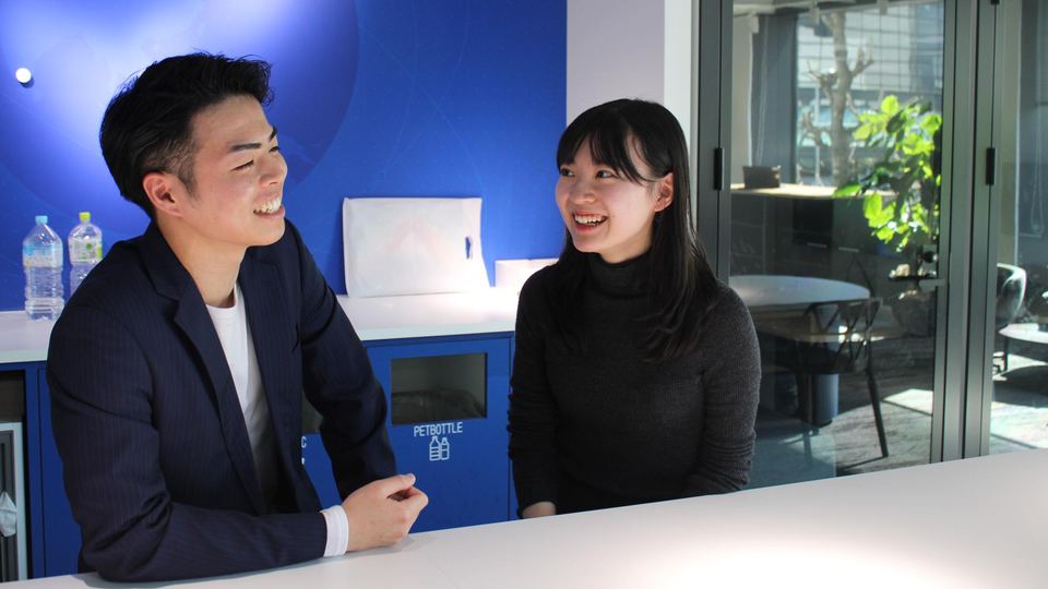 オフィスの青い壁の前で笑いながら話すメンバー2人